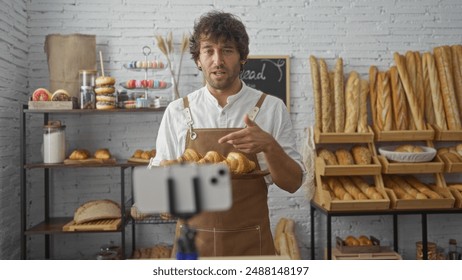 Young, hispanic, man in a bakery holding baked goods, surrounded by various breads and pastries, with a smartphone on a tripod capturing the scene in an indoor shop setting. - Powered by Shutterstock