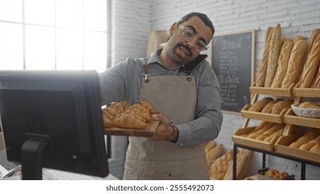 Young hispanic man in bakery shop holds tray of croissants while on phone call indoors - Powered by Shutterstock