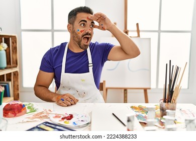 Young hispanic man and beard at art studio and painted face very happy   smiling looking far away and hand over head  searching concept  