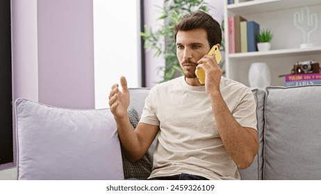 A young hispanic man with a beard engages in conversation on a yellow phone while sitting on a grey couch indoors. - Powered by Shutterstock