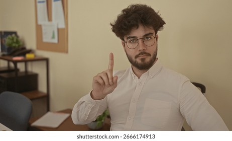 Young hispanic man with beard gesturing no in an office setting, wearing glasses and a button-up shirt while sitting indoors with bulletin board and papers in background.