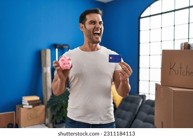 Young hispanic man with beard holding piggy bank and credit card at new home angry and mad screaming frustrated and furious, shouting with anger. rage and aggressive concept.  - Shutterstock ID 2311184113