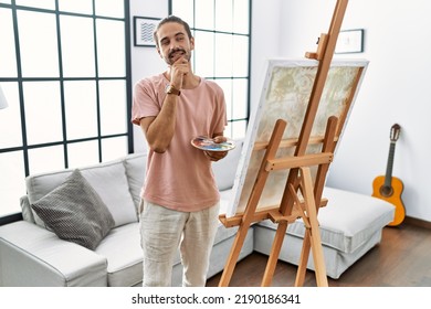 Young Hispanic Man With Beard Painting On Canvas At Home With Hand On Chin Thinking About Question, Pensive Expression. Smiling And Thoughtful Face. Doubt Concept. 