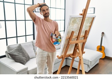 Young Hispanic Man With Beard Painting On Canvas At Home Confuse And Wondering About Question. Uncertain With Doubt, Thinking With Hand On Head. Pensive Concept. 