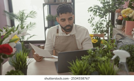 A young hispanic man with a beard reviews a document at a flower shop's indoor workspace surrounded by fresh plants. - Powered by Shutterstock