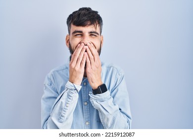 Young Hispanic Man With Beard Standing Over Blue Background Laughing And Embarrassed Giggle Covering Mouth With Hands, Gossip And Scandal Concept 