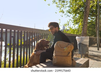 Young Hispanic Man With Beard, Sunglasses, Black Shirt And Backpack, Sitting On A Bench Petting His Dog Under The Sun Rays. Concept Animals, Dogs, Love, Pets, Golden.