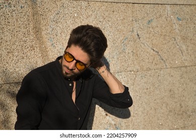Young Hispanic man with a beard, sunglasses and black shirt, touching the back of his neck, sorry, afflicted, sad, leaning on a stone wall. Concept of grief, loneliness, disappointment, expressions. - Shutterstock ID 2204257909