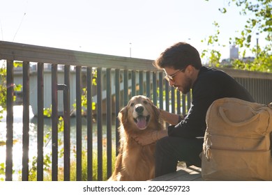 Young Hispanic Man With Beard, Sunglasses, Black Shirt And Backpack, Sitting On A Bench Petting His Dog Under The Sun Rays. Concept Animals, Dogs, Love, Pets, Golden.