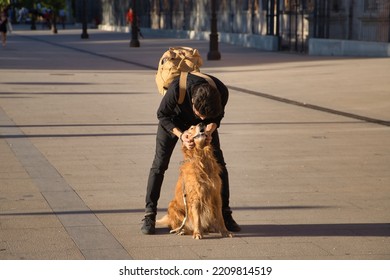 Young Hispanic Man With Beard, Sunglasses, Black Shirt And Backpack, Crouched Hugging His Dog In The City. Concept Animals, Dogs, Love, Pets, Golden.