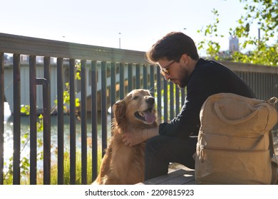 Young Hispanic Man With Beard, Sunglasses, Black Shirt And Backpack, Sitting On A Bench Petting His Dog Under The Sun Rays. Concept Animals, Dogs, Love, Pets, Golden.