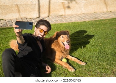 Young Hispanic Man With A Beard, Sunglasses And Black Shirt, Taking A Selfie With His Mobile, Lying On The Lawn Next To His Dog Sunbathing. Concept Animals, Dogs, Love, Pets, Gold.