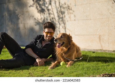 Young Hispanic Man With A Beard, Sunglasses And Black Shirt, Lying On The Lawn Next To His Dog Sunbathing. Concept Animals, Dogs, Love, Pets, Gold.