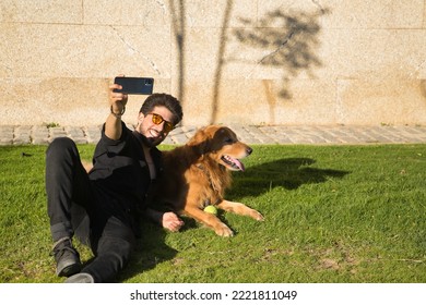 Young Hispanic Man With A Beard, Sunglasses And Black Shirt, Taking A Selfie With His Mobile, Lying On The Lawn Next To His Dog Sunbathing. Concept Animals, Dogs, Love, Pets, Gold.