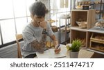 Young, hispanic, man with a beard and tattoos sits pensively in a bakery, writing in a notebook with shelves of bread and pastries in the background