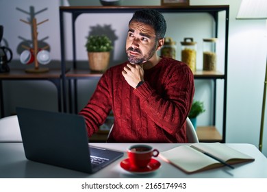 Young Hispanic Man With Beard Using Computer Laptop At Night At Home With Hand On Chin Thinking About Question, Pensive Expression. Smiling With Thoughtful Face. Doubt Concept. 