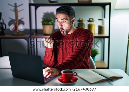 Young hispanic man with beard using computer laptop at night at home surprised pointing with hand finger to the side, open mouth amazed expression. 