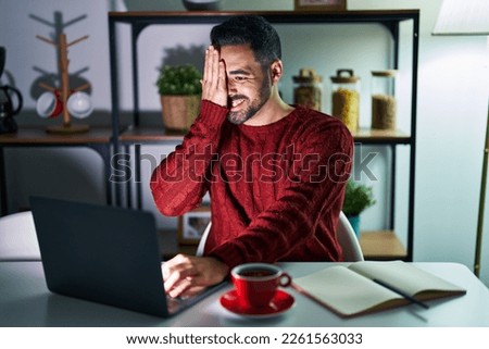 Young hispanic man with beard using computer laptop at night at home covering one eye with hand, confident smile on face and surprise emotion. 