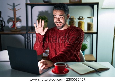 Young hispanic man with beard using computer laptop at night at home waiving saying hello happy and smiling, friendly welcome gesture 