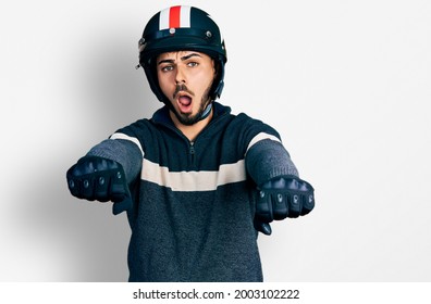 Young Hispanic Man With Beard Wearing Motorcycle Helmet Doing Driving Gesture In Shock Face, Looking Skeptical And Sarcastic, Surprised With Open Mouth 