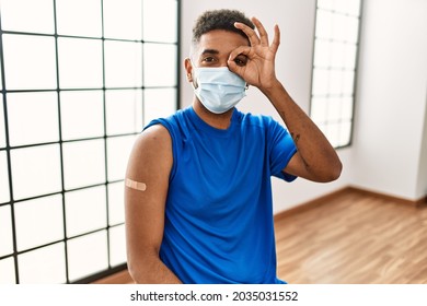 Young Hispanic Man With Beard Wearing Safety Mask Getting Vaccine Smiling Happy Doing Ok Sign With Hand On Eye Looking Through Fingers 