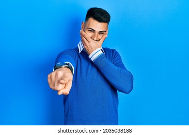 Young Hispanic Man With Beard Wearing Casual Blue Sweater Laughing At You, Pointing Finger To The Camera With Hand Over Mouth, Shame Expression 