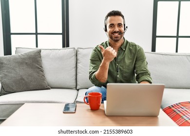 Young Hispanic Man With Beard Wearing Call Center Agent Headset Working From Home Pointing To The Back Behind With Hand And Thumbs Up, Smiling Confident 