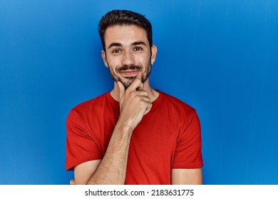 Young Hispanic Man With Beard Wearing Red T Shirt Over Blue Background Looking Confident At The Camera Smiling With Crossed Arms And Hand Raised On Chin. Thinking Positive. 