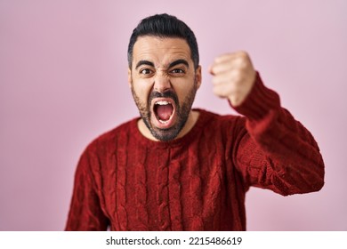 Young hispanic man with beard wearing casual sweater over pink background angry and mad raising fist frustrated and furious while shouting with anger. rage and aggressive concept.  - Shutterstock ID 2215486619