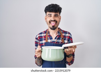 Young Hispanic Man With Beard Wearing Apron Holding Cooking Pot Sticking Tongue Out Happy With Funny Expression. 