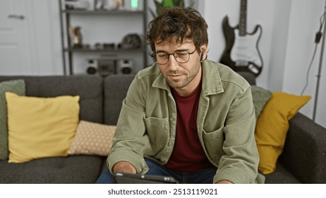 A young hispanic man with a beard wearing wireless earbuds and glasses, focused on a tablet indoors. - Powered by Shutterstock