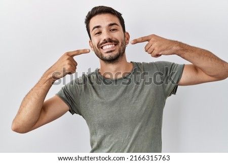 Young hispanic man with beard wearing casual t shirt over white background smiling cheerful showing and pointing with fingers teeth and mouth. dental health concept. 