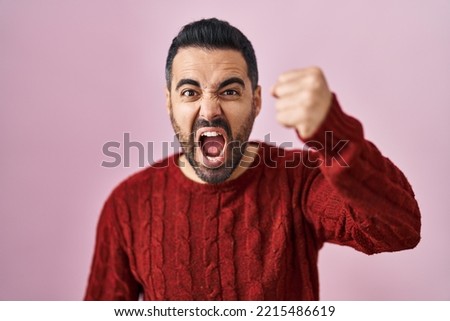 Young hispanic man with beard wearing casual sweater over pink background angry and mad raising fist frustrated and furious while shouting with anger. rage and aggressive concept. 