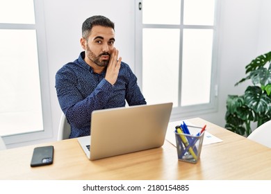 Young Hispanic Man With Beard Working At The Office With Laptop Hand On Mouth Telling Secret Rumor, Whispering Malicious Talk Conversation 