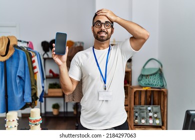 Young hispanic man with beard working as manager at retail boutique holding smartphone stressed and frustrated with hand on head, surprised and angry face 