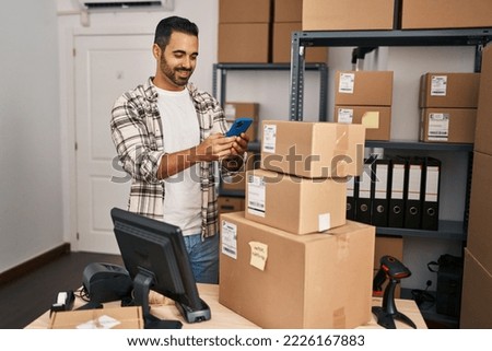 Young hispanic man with beard working at small business ecommerce with smartphone looking positive and happy standing and smiling with a confident smile showing teeth 