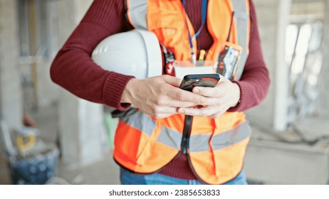 Young hispanic man builder using smartphone holding hardhat at construction site