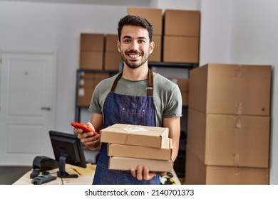 Young hispanic man business worker using smartphone holding packages at storehouse - Shutterstock ID 2140701373
