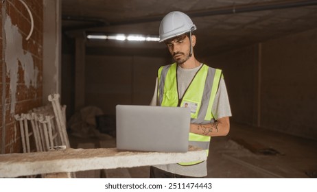 Young hispanic man at a construction site wearing safety gear and using a laptop with a focused expression - Powered by Shutterstock