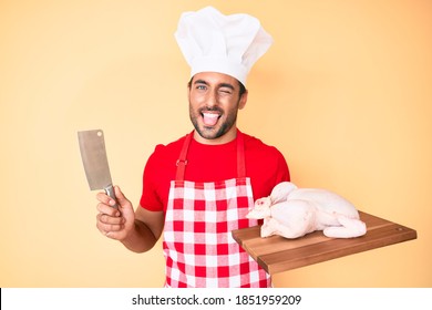 Young Hispanic Man Cooking Chicken Holding Knife Sticking Tongue Out Happy With Funny Expression. 