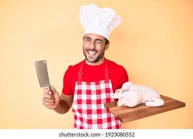 Young Hispanic Man Cooking Chicken Holding Knife Winking Looking At The Camera With Sexy Expression, Cheerful And Happy Face. 