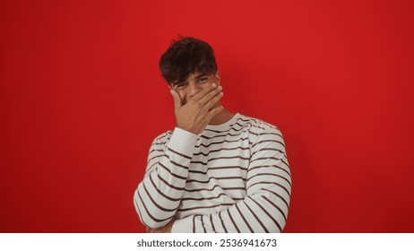 A young hispanic man covers his mouth with his hand while standing against a vibrant red background, wearing a striped sweater. - Powered by Shutterstock