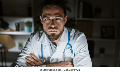 Young hispanic man doctor sitting on table with arms crossed gesture and serious face at the clinic - Powered by Shutterstock