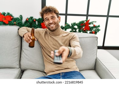Young Hispanic Man Drinking Beer Watching Tv Sitting By Christmas Decor At Home