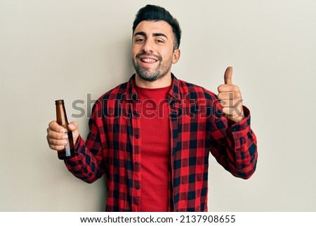 Young hispanic man drinking a bottle of beer smiling happy and positive, thumb up doing excellent and approval sign 