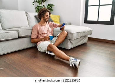 Young Hispanic Man Drinking Coffee And Using Smartphone Sitting On The Floor At Home.