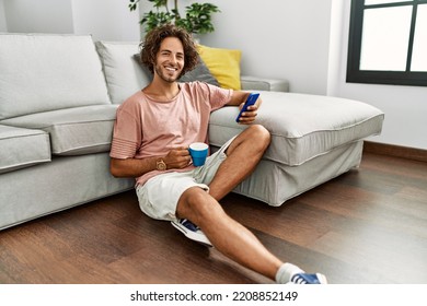 Young hispanic man drinking coffee and using smartphone sitting on the floor at home. - Powered by Shutterstock