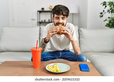 Young Hispanic Man Eating Classical Burger And Drinking Soda Sitting On The Sofa At Home.