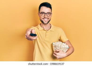 Young hispanic man eating popcorn using tv control smiling with a happy and cool smile on face. showing teeth. 