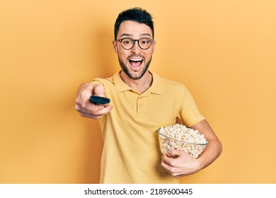 Young hispanic man eating popcorn using tv control smiling and laughing hard out loud because funny crazy joke.  - Powered by Shutterstock